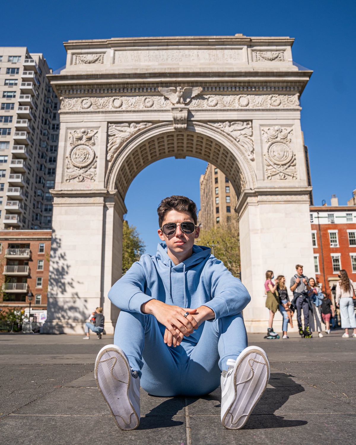 Eitan Bernath sits in blue pastel sweatsuit in front of Washington Arch in the park in NYC.