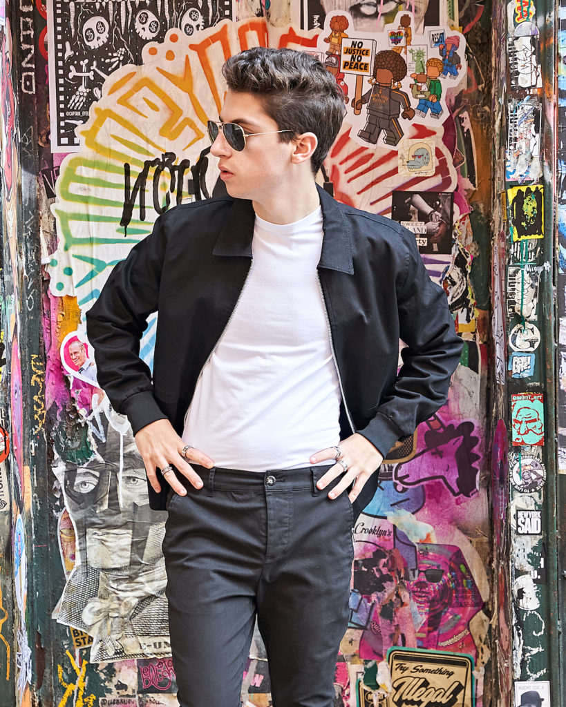 Eitan Bernath wears white tee shirt and black jacket in front of graffiti wall in NYC.