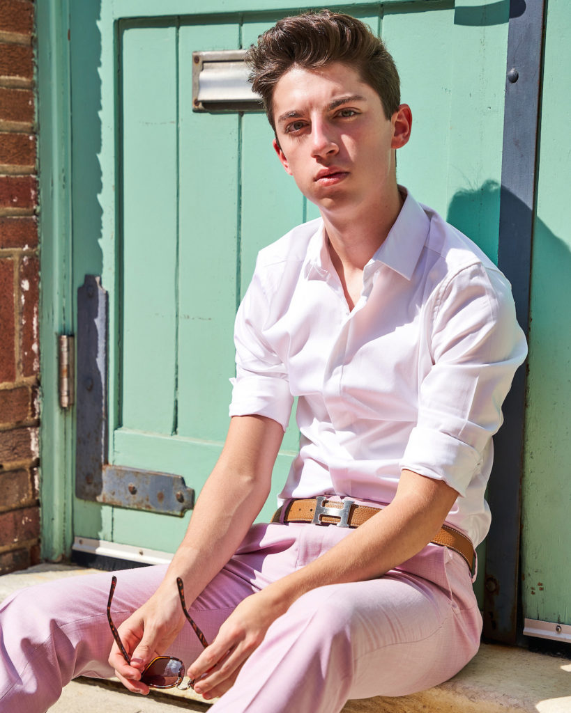 Eitan Bernath wears pastel pants and sits on the street in front of a green door in NYC.